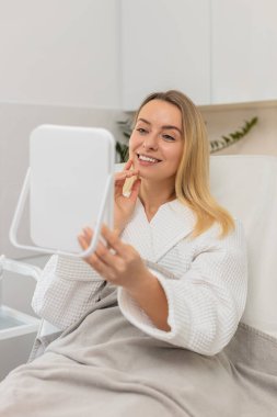 Beyaz kadın güzellik kliniğinde oturuyor ve kozmetoloji ameliyatından sonra aynada yüzünü kontrol ederken gülümsüyor. Orta yaşlı bayan tedaviden memnun, sağlıklı cilt sonuçlarının tadını çıkarıyor..