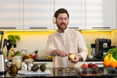 Orta yaşlı bir adam evde elma soyarken sağlıklı atıştırmalıklar ve diyet planı için gülümsüyor. Genç adam, sabah yemeği hazırlığı sırasında evde temiz beslenme için taze meyve hazırlıyor.