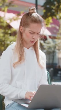 Laptop kullanan, kameraya bakan ve şehir sokaklarında sandalyeye otururken baş parmağını öne eğen genç bir kadın. Kafkas kız olumsuz duygularını ifade eden bir defter üzerinde çalışıyor..