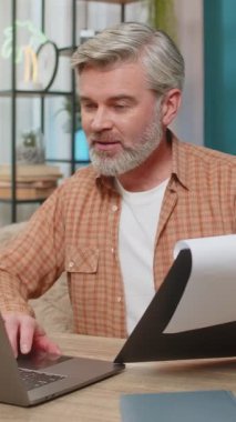 Beyaz, olgun bir adam evde oturmuş önemli belgeleri inceliyor ve dizüstü bilgisayar kullanıyor. Orta yaşlı bir adam başarılı bir işten aldığı memnuniyetle gülümser, verimlilik ve başarıyı gösterir.