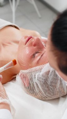Kozmetik uzmanı klinikte yüz fırçasıyla yüz maskesi takarken beyaz bir kadın kanepede yatıyor. Rahatlama, cilt bakımı, pürüzsüz cilt rahatlığı, sağIık ve seans sırasında sakin bir ruh hali verir.