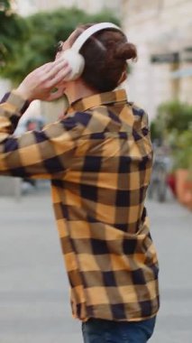 Kablosuz kulaklıklı mutlu turistin arka görüntüsü açık havada akıllı telefondan en sevilen enerjik disko müziğini dinliyor. Beyaz tenli turist, şehrin güneşli sokağından geçiyor..
