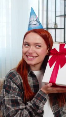 Evde parti şapkası takan genç bir kadın güzel bir sürpriz tepkiyle doğum günü hediyesi veriyor ve gülümsüyor. Kafkasyalı kızıl saçlı kız kanepenin üzerinde sevinç ve duygusal zevkle kutlama anının tadını çıkarıyor.