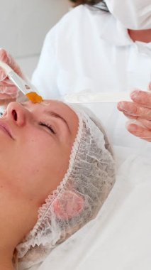 Kozmetik bilimci kozmetoloji kliniğinde yüz maskesi takarken beyaz bir kadın kanepede uzanıyor. Rahatlama cilt bakımı, pürüzsüz cilt konforu sağIık ve sakin kaplıca havası verir.