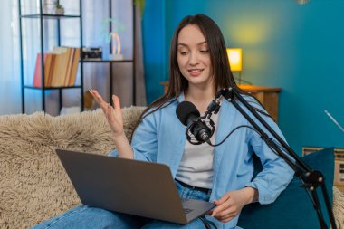 Genç kadın blogcu mutlu bir şekilde podcast veya vlog kaydediyor dizüstü bilgisayarı ve stüdyo mikrofonuyla kanepede oturuyor, hareketli bir şekilde. İnternette duygusal açıdan ilgi çekici, ilginç bir hikaye anlatan beyaz bir kız.