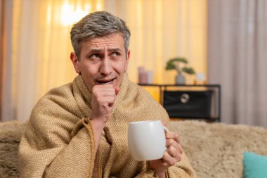 Evdeki üzgün genç adam bir fincan çayla battaniyeye sarılmış, hasta görünüyor ve öksürüyor. Kanepedeki beyaz adam soğuk algınlığı belirtileri gösteriyor, sessizce dinleniyor ve hafif bir hastalık ve yorgunluktan kurtulmaya çalışıyor.