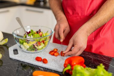 Arap adam kiraz domateslerini dilimliyor ve sağlıklı beslenme ve kilo kontrolü için mutfağa sebze ekliyor. Genç adam evde yemek hazırlamaya odaklanıyor. Temiz yemek ve sağlıklı yaşam için..