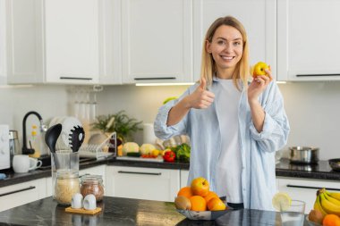 Orta yaşlı bir kadın mutfak masasında taze elma yiyor. Vitamin ve diyet için sağlıklı atıştırmalıkların tadını çıkarıyor. Genç kız, günlük temiz yemek ve sağlık rutinini destekleyen tatlılar yerine meyve seçiyor..