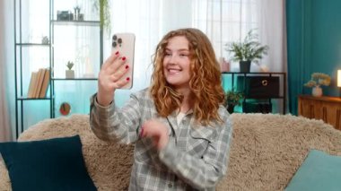 Genç bir kadın akıllı telefonuyla arkadaşını sevinçle karşılıyor ve mutlu haberi paylaşıyor. Kafkasyalı kızıl kız kanepede dinleniyor, neşeli bir şekilde gülümsüyor, pozitif etkileşim içinde..