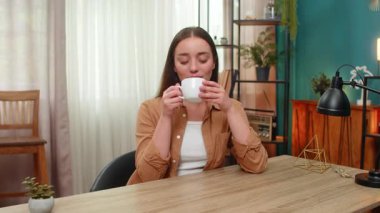 Masadaki genç kadın kahve fincanını kaldırıyor ve hafifçe gülümsüyor dengeli sakinliğin ve sessizliğin tadını çıkarıyor. Kafkasyalı kız yumuşak sıcaklığın tadını alıyor, huzurlu düşüncelerin sessiz bir minnettarlıkla dolaşmasına izin veriyor.