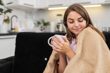 Evdeki genç kadın bitkisel çay içiyor ve kupanın üzerinde ellerini ısıtıyor ve yavaş yavaş nefes alıyor ve sakinleşiyor. Kanepede oturan beyaz kız gülümsüyor ve sessizliğin tadını çıkarıyor ve dinleniyor ve sıcak bir ruh hali içinde dinleniyor ve rahatlıyor.