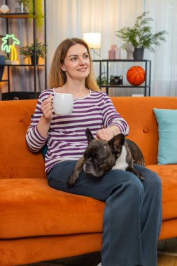 Evde Fransız bulldog köpeği olan orta yaşlı bir kadın sabah çayını yudumlar ve sakinleşir. Kanepede köpekli bir kız gülümseyerek sıcak çay fincanının tadını çıkarıyor.