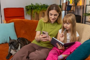 Evde Fransız bulldog 'u olan anne ve kızı matematik dersi için akıllı telefon kullanıyor. Kanepede köpeği olan kadın ve kız iş çözer. Yaşasın gülümseme. Keyifle öğren.