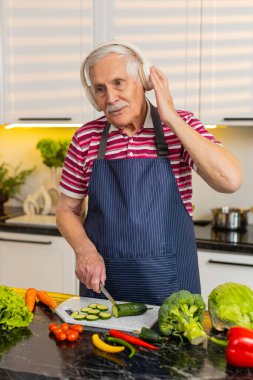 Yaşlı adam evde kulaklık takarken, diyet salatası için gülümseyip sebze dilimliyor. Yaşlı büyükbaba, evde kilo kaybı ve sağIık için müzik ve sağlıklı yemek yapmayı sever..