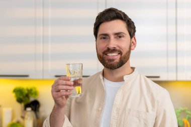 Orta yaşlı bir adam sabah rutini sırasında kendini dinlenmiş ve sakin hissederek mutfakta camdan su içer. Genç adam sağlıklı diyet sağlık ve kilo verme hedefleri için hidrasyon alışkanlığını destekliyor..