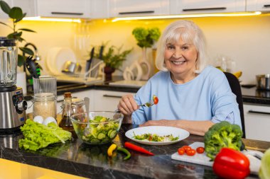 Diyet desteği için ev mutfağında taze sebze salatası yerken gülümseyen yaşlı bir kadın. Sağlıklı yemek seçiminin tadını çıkaran yaşlı bir büyükanne. Sağlıklı beslenmek için sakin bir ortam..