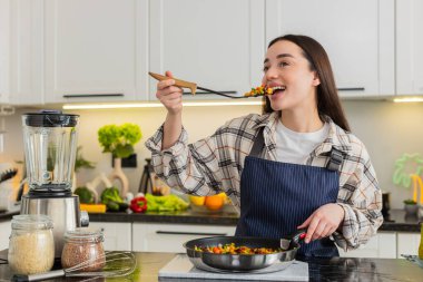 Genç kadın, diyet planı ve günlük sağlık için evde yemek pişirirken tavadaki sebzeleri tadıyor. Tat testinden sonra kız tatmin olmuş görünüyor. Ev yapımı öğle yemeğini evde servis etmeye hazır..