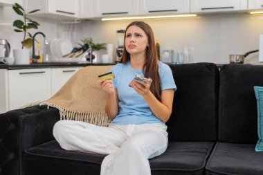 Evdeki genç kadın akıllı telefondan ödemeyi kredi kartıyla yapmaya çalışıyor. Düşük bütçeli ve engelli olduğunu düşünüyor. Kanepedeki beyaz kız dolandırıcılık uyarısı okuyor. Para kaybından korkuyor ve banka desteğini arıyor..