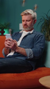 Mature senior bearded man lying on sofa uses mobile phone smiles at night evening home. Mature guy texting share messages content on smartphone social media applications online watching relax movie.