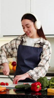 Genç bir kadın mutfakta vejetaryen salatası için vişneli domatesleri kesip kulaklıkla müzik dinliyor. Kız evde, evde sağlıklı yiyecekler hazırlarken rahat kalıyor. Diyet için.