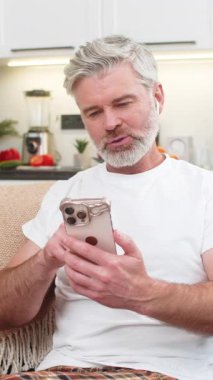 Evde orta yaşlı bir adam akıllı telefon uygulamasından kelimeleri kulaklıkla öğreniyor ve geri bildirim duyuyor. Olgun adam koltukta durur ve gülümser. Yüksek skoru görür. Büyük ilerleme kaydeder ve çalışmaya devam eder..