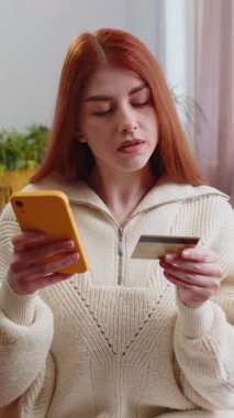 Genç kızıl saçlı bir kadın kredi kartı ve akıllı telefon kullanarak para aktarıyor, internetten nakit para almıyor, ev içinde yemek siparişi veriyor. Mutlu bayan kanepede oturuyor.