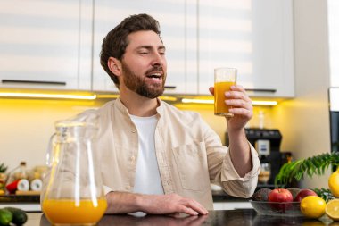 Orta yaşlı bir adam mutfakta taze portakal suyu içiyor sağlıklı beslenme ve su ihtiyacını gidermek için vitamin takviyesi yapıyor. Genç adam spor yaptıktan sonra evde rahatlıyor. Sağlık kilo kaybı ve beslenme hedeflerini destekliyor..