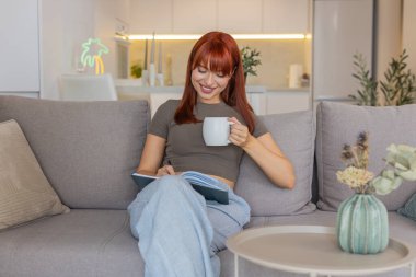 Evde sıcak çay içen genç bir kadın nefes alıyor, zihni yavaşlatıyor ve ertesi gün not defterine kontrol listesi yazıyor. Kanepedeki kızıl saçlı kız, yarın için sakin bir odak planı buldu. Sıcak bir fincan ellerde ve yumuşak bir barış.