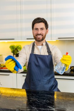 Orta yaşlı bir adam mutfak tezgahını bezle ve temizlik spreyiyle siliyor. Evde temizlik ve gıda güvenliği için eldiven giyiyor. Genç adam, modern mutfaktaki hafta sonu işlerinden sonra mutlu ve tatmin olmuş hissediyor..
