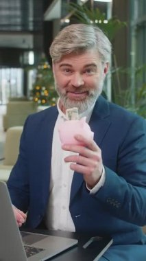 Olgun iş adamı, pembe kumbarayla para biriktirir ve içine dolar atar. Başarı, ekonomi, yatırım, depozito. Modern ofis lobisinde dizüstü bilgisayarlı takım elbiseli adam oturuyor..