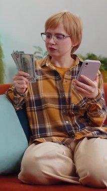 Aile bütçesi planlamak. Gülümseyen genç bir kadın nakit para sayıyor, akıllı telefon kullanıyor, ev faturalarını hesaplıyor. Gözlüklü sarışın kadın kanepede oturur. Oturma odasındaki maaşından memnun kalır.