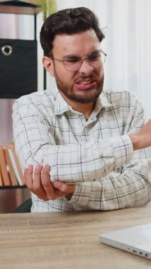 Yorgun Hintli Arap iş adamı yönetici ev ofisinin masasında dizüstü bilgisayarla oturuyor ve dirsek ekleminde ağrı ve rahatsızlık hissediyor ve acı verici bir el masajı yapıyor. Eklem iltihabı ve kemik iltihabı kavramları.