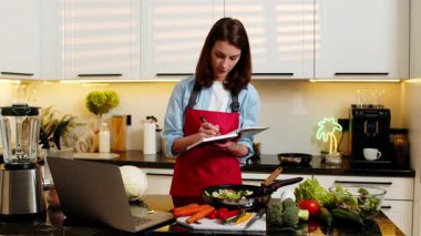 Genç bir kadın ev mutfağında sağlıklı yemek hazırlamak için not alırken dizüstü bilgisayarında dengeli bir diyet tarifi uyguluyor. Yetişkin kız evde kilo verme yaşam tarzı için beslenme planına odaklanmış olmaktan mutlu..