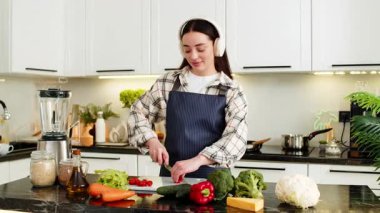 Genç bir kadın, temiz beslenme için evde vejetaryen salatası yapan kulaklık takan domatesleri kesiyor. Kız evde sakin bir şekilde sağlıklı yemek hazırlarken müziği seviyor..