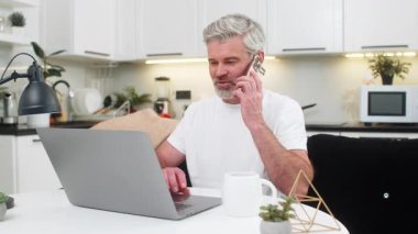 Evde dizüstü bilgisayarı olan orta yaşlı bir adam akıllı telefondan müşteri sesiyle sakin bir şekilde arıyor. Masada oturan olgun adam güncellemeleri yazar soruları cevaplar ve hedeflere ulaşırken iş akışını pürüzsüz tutar