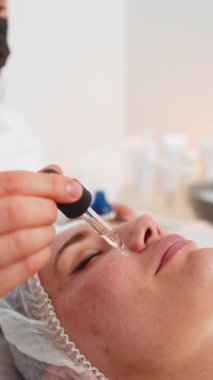 Orta yaşlı yetişkin bir kadın kozmetoloji kliniğinde cilt bakımı sırasında yüz serumu alır. Güzellik uzmanı su, esneklik, ışıltı, beslenme ve görünür gençleşme için pipetle sıvı uyguluyor.