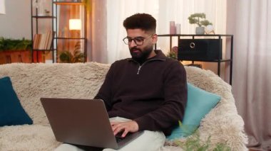 Evdeki Hintli adam online sınav sonuçlarını görüyor ve geniş bir gülümseme sergiliyor, başarı üstüne sevinçle yumruğunu kaldırıyor. Kanepedeki Arap adam başarıya sevinçle tepki veriyor, gururlu ve pozitif duygularla heyecanlanıyor.