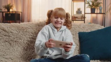 Genç kız evde akıllı telefon kullanıyor, oyun oynarken kıkırdıyor ve heyecanla oyun seviyesini geçiyor. Çocuk koltukta elinde telefonla zaferi kutluyor. Neşeli bir zafer.