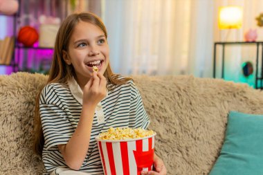 Genç kız patlamış mısır yer ve kanepede oturup eğlenirken televizyonda komik çizgi filmler izler. Okullu çocuk evde rahat bir film izlerken keyifli ve rahat görünüyor.