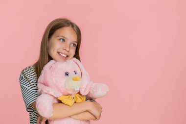 Genç kız pelüş oyuncak tavşancığa sevgi ve mutlulukla sarılıyor, sevinçle tatlı tatlı gülümsüyor, sıcaklığını ve duygusal bağını ifade ediyor. Pembe arka planda izole edilmiş küçük okul çocuğu..