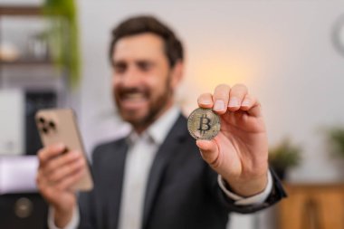 Merkez ofisteki orta yaşlı işadamı akıllı telefondan Bitcoin tablosunu izliyor. Masadaki serbest çalışan adam büyük kripto zenginlik hedeflerini kutluyor..