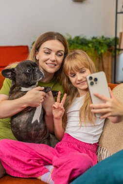 Evde Fransız bulldog 'u olan anne ve kız selfie çekiyor. Akıllı telefon maskesi filtresi takıp gülüyor ve sırıtıyor. Kanepede köpekli bir kadın ve kız kamerayla oynuyorlar. Fotoğraf çekiniyorlar. Eğlenceli aile anıları paylaşıyorlar.