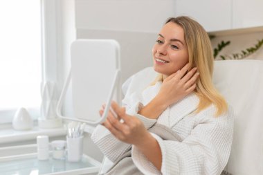 Beyaz kadın güzellik kliniğinde oturuyor ve kozmetoloji ameliyatından sonra aynada yüzünü kontrol ederken gülümsüyor. Orta yaşlı bayan tedaviden memnun, sağlıklı cilt sonuçlarının tadını çıkarıyor..