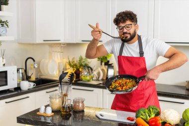 Arap adam tavada sebze karıştırır ve mutfakta sağlıklı bir akşam yemeği için kaşık dolusu lezzet kontrolü yapar. Genç adam evde yemek pişirmeyi, diyet yemek tarifini evde evde öğrenmeyi seviyor..