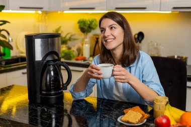 Genç bir kadın, ev mutfağında taze demlenmiş kahve aromasını sever. Huzurlu bir sabah kahvaltısı sırasında sıcak mutluluğu hisseder. Yetişkin kız, diyet hedefleri için sağlıklı yaşam tarzını destekleyen sıcak bir bardakla rahatlıyor..