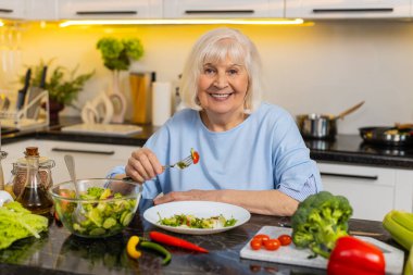 Diyet desteği için ev mutfağında taze sebze salatası yerken gülümseyen yaşlı bir kadın. Sağlıklı yemek seçiminin tadını çıkaran yaşlı bir büyükanne. Sağlıklı beslenmek için sakin bir ortam..