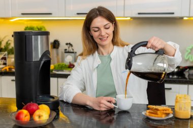 Orta yaşlı bir kadın kahvaltı ve enerji takviyesi için ev mutfağında kahvesini fincana doldurur. Genç yetişkin bir kız sabah fıstık ezmeli tost içerken gülümser ve internette sağlıklı beslenme planları yapar..