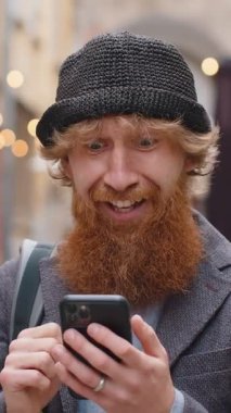 Genç sakallı adam cep telefonuyla kutlama yapıyor. İyi haber kazan, piyango kazan, internetten bağış yap. Şehir sokaklarında gezen mutlu kızıl saçlı çocuk. Yaşam Tarzları