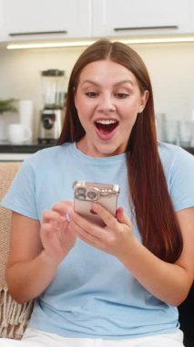 Akıllı telefondaki finans uygulamasını kontrol eden genç bir kadın dengenin arttığını ve kazandığını kutluyor. Kanepede oturan beyaz kız online oyundan sonra ikramiye için yumruğunu kaldırıyor. Cep telefonu uygulamasında kazanıyor. Çok heyecanlı..