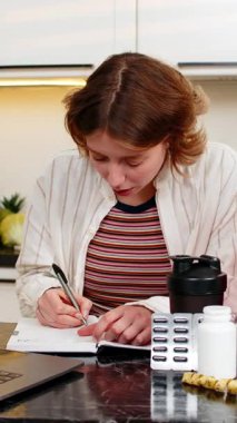 Diyet kontrolü ve egzersiz hedefleri için not defterine form takviyesi yazarken gülümseyen genç bir kadın. Yetişkin kadın, sağlıklı ev planlaması sırasında düzenli eğitim için dikkatli beslenme seçimleri yapar.